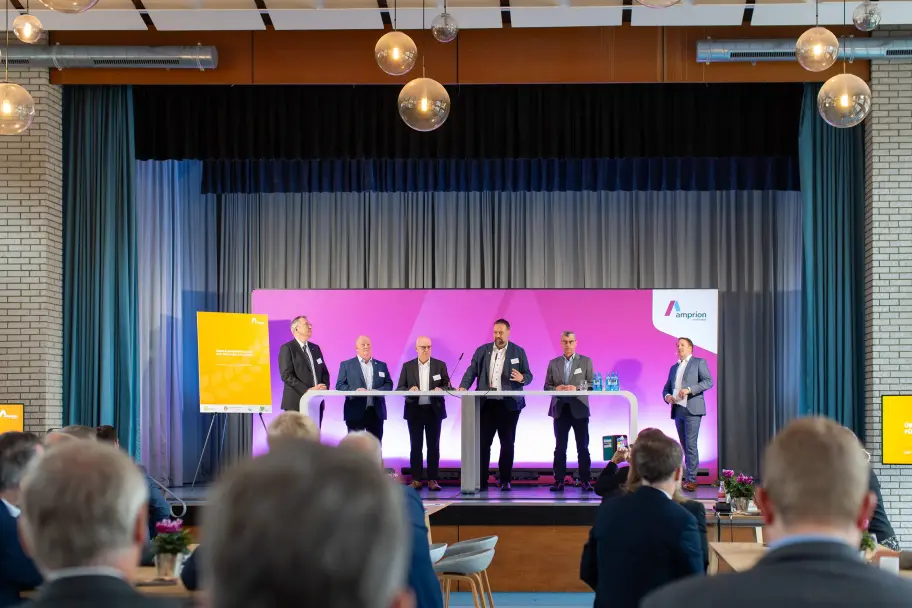 Podiumsdiskussion auf einer Bühne mit sieben Männern in Anzügen, die hinter einem weißen Stehtisch stehen. Im Hintergrund ein magentafarbener Hintergrund mit Amprion-Logo und ein gelbes Plakat mit der Aufschrift „Über-Rahmenregelung für Erdkabelprojekte 2025“. Vor der Bühne sitzt das Publikum.