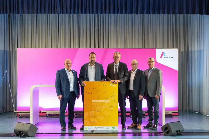 Fünf Männer in Anzügen stehen auf einer Bühne und halten gemeinsam ein gelbes Plakat mit Unterschriften, das die neue Rahmenregelung für Erdkabelprojekte 2025 zeigt. Der Hintergrund ist magentafarben mit Amprion-Logo.