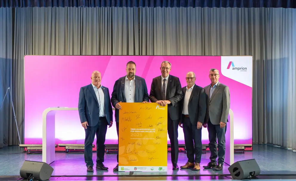 Fünf Männer in Anzügen stehen auf einer Bühne und halten gemeinsam ein gelbes Plakat mit Unterschriften, das die neue Rahmenregelung für Erdkabelprojekte 2025 zeigt. Der Hintergrund ist magentafarben mit Amprion-Logo.
