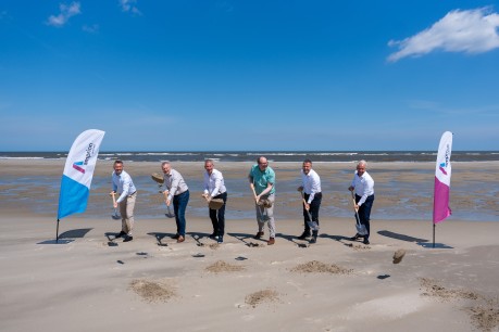 Sechs Männer in Hemden und Hosen stehen nebeneinander auf einem Sandstrand und halten Spaten in den Händen, als würden sie etwas ausgraben. Sie blicken alle in Richtung des Betrachters. Hinter ihnen erstreckt sich das Meer unter einem klaren blauen Himmel mit wenigen Wolken. Links und rechts im Bild sind jeweils eine Fahne mit dem Logo "amprion" zu sehen. Die Fahne auf der linken Seite ist blau und weiß, die auf der rechten Seite ist violett und weiß.